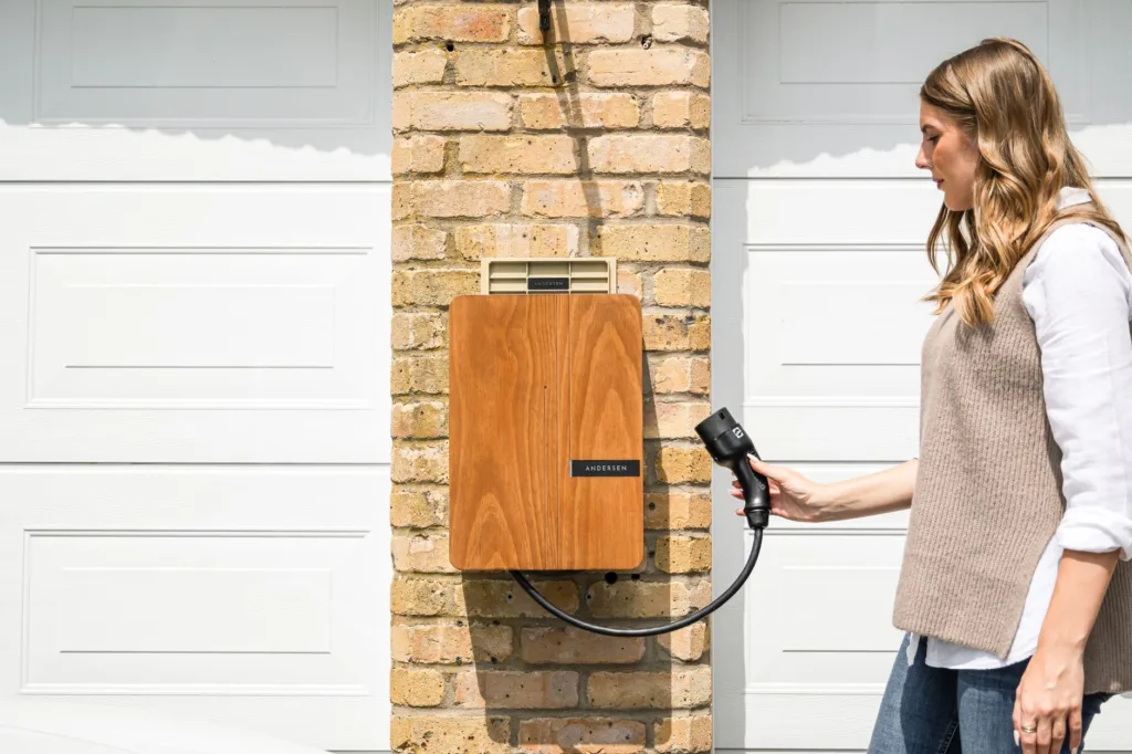 Wall-mounted Andersen EV charger with wooden cover on brick pillar between garage doors, woman holding charging plug