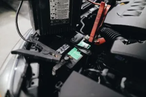 Car battery with red jumper clamp and portable jump starter attached under hood, showing jumper cables ready for road trips