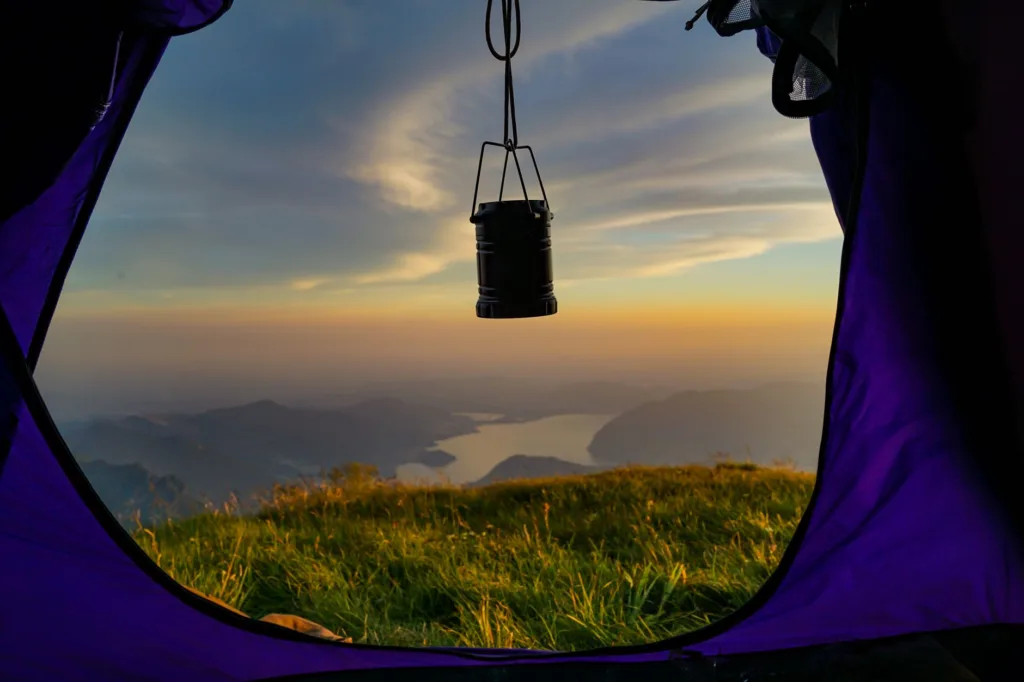 Silhouetted LED lantern hanging in tent doorway at sunset, overlooking grassy hill and lake — camping lantern scene