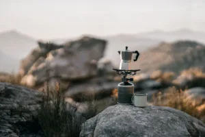 Moka pot on a portable camping gas stove with enamel mug on rock, compact portable coffee maker for camping in the mountains