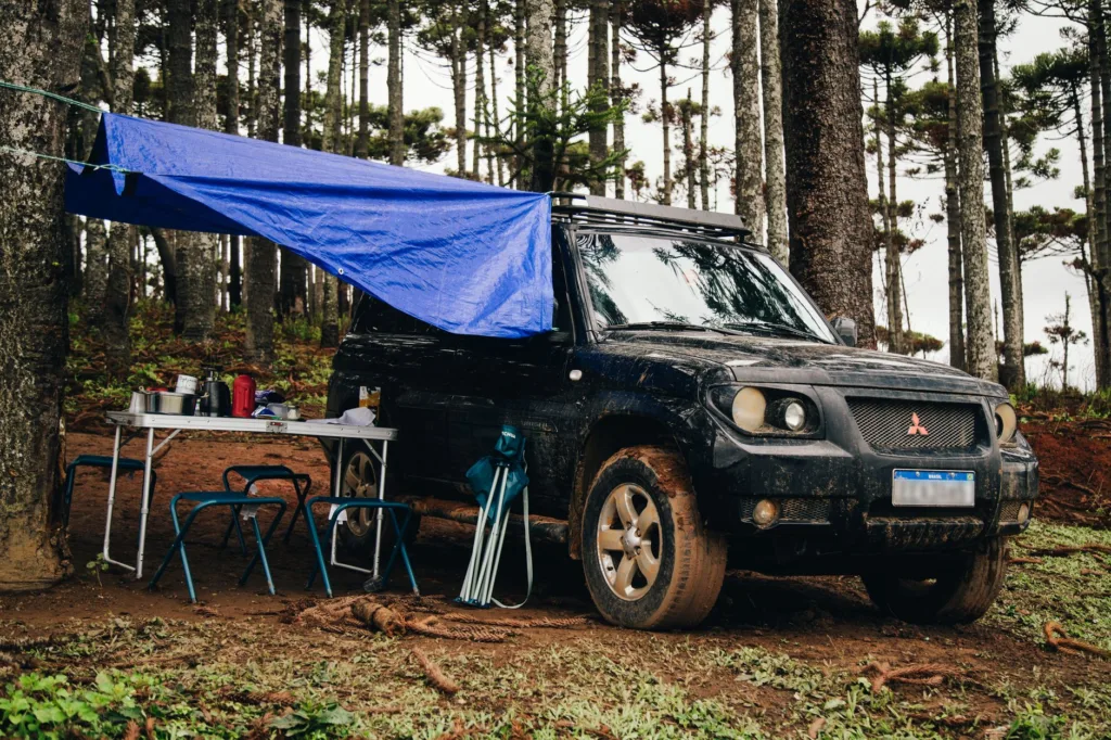 Muddy SUV in forest with blue tarp awning, folding table and chairs, showcasing privacy curtains for SUV camping