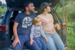 Family of three sitting in open hatchback trunk, enjoying countryside view during a family road trip with packed essentials.