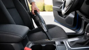 Handheld car vacuum cleaning crumbs from a dark car seat beside the center console